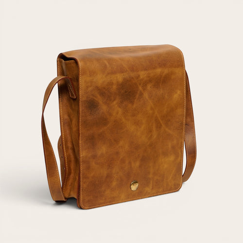 Bandolera vertical de cuero genuino OLKI en tono habano rústico con solapa y broche metálico.
