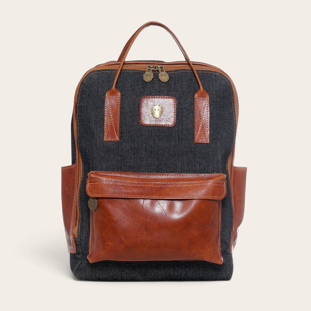 Mochila OLKI Caliboro de mezclilla gris con detalles de cuero marrón, bolsillo frontal y asas superiores.